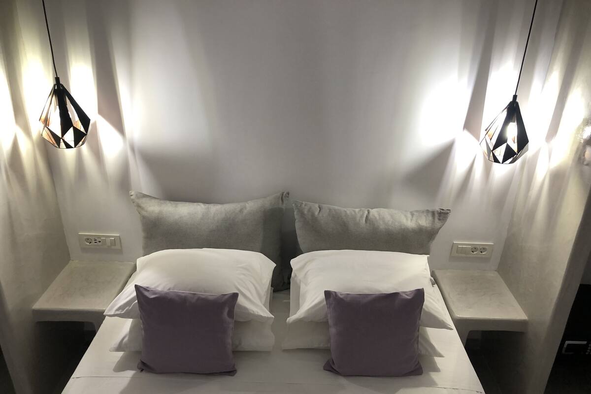 A room with two beds, each with a pillow and a nightstand. There are two pendant lights hanging from the ceiling.