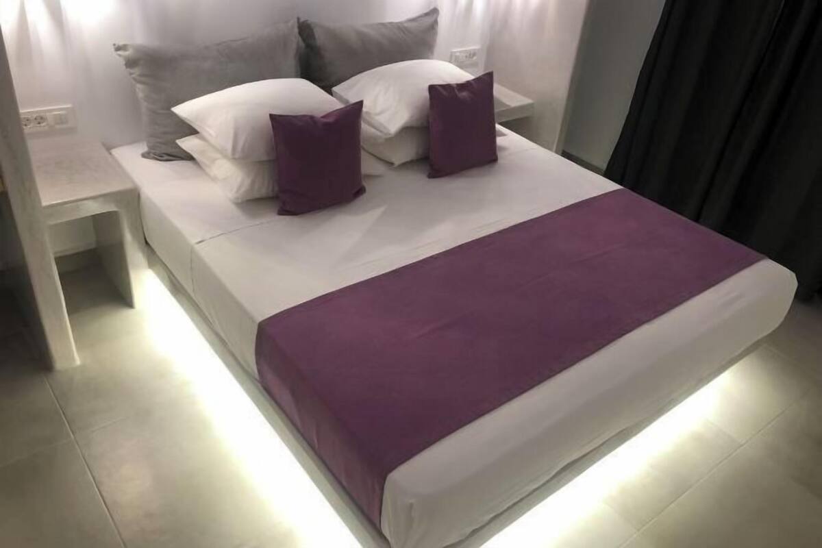 A neatly made bed with a white mattress and a purple blanket, flanked by two white pillows and two purple accent pillows.