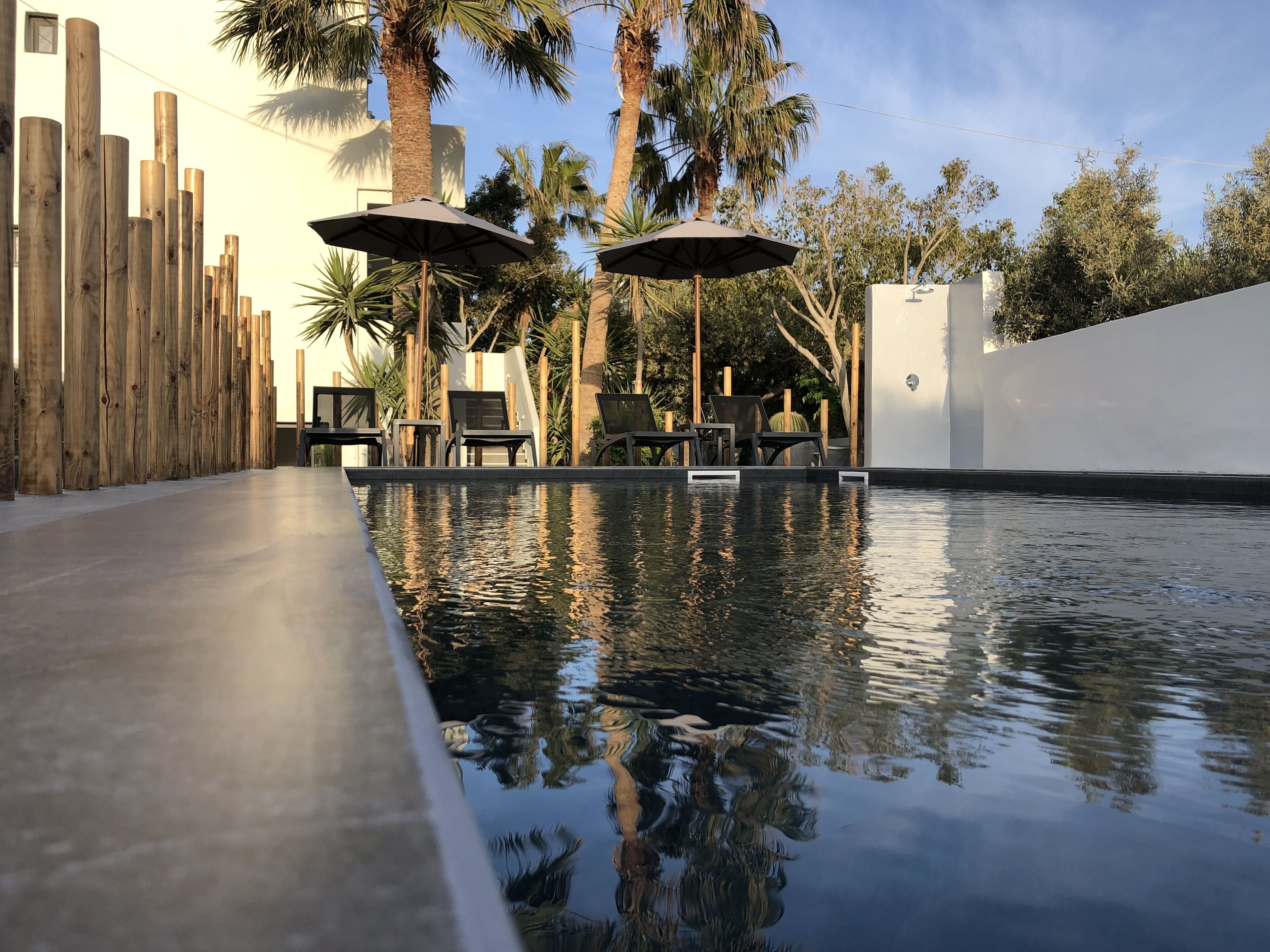 A swimming pool with lounge chairs and umbrellas, surrounded by wooden pillars and palm trees.