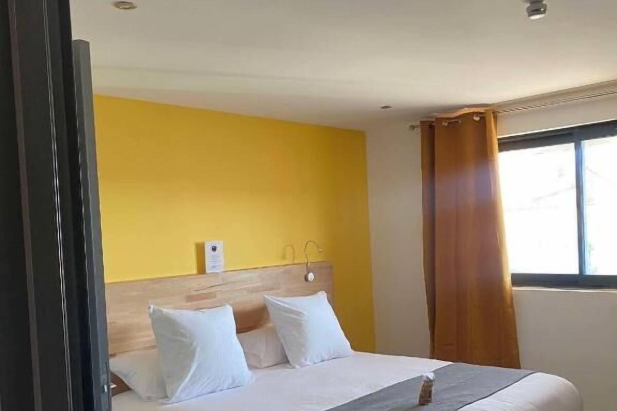 A hotel room with a bed, a wooden headboard, a window with curtains, and a yellow wall.