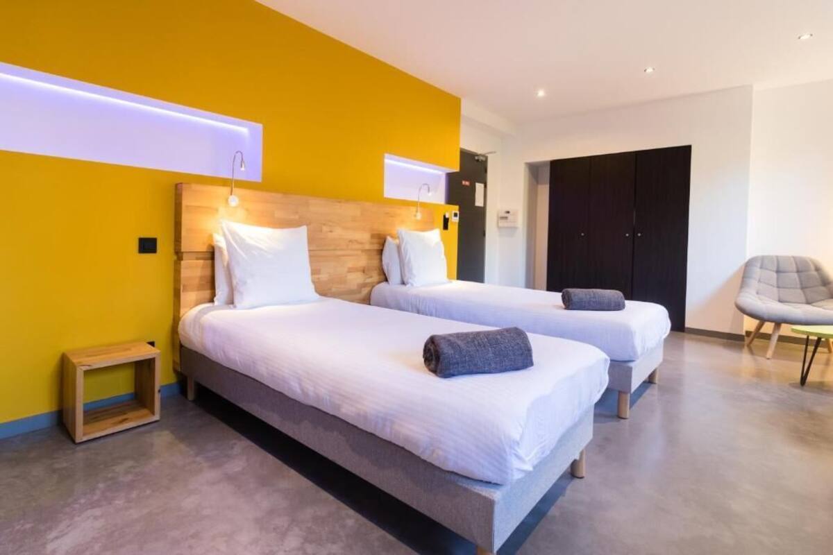 A hotel room with two beds, a wooden headboard, a small wooden stool, and a grey armchair.