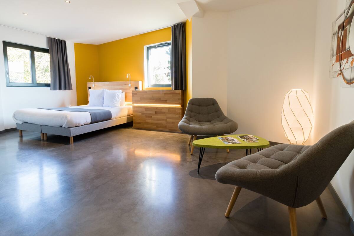 A modern hotel room with a bed, two armchairs, a small table, and a window with curtains.