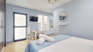 A bedroom with a bed, a desk, a chair, a TV, and a large window. - Eden on the Bay Apartment 67B (Cape Town)