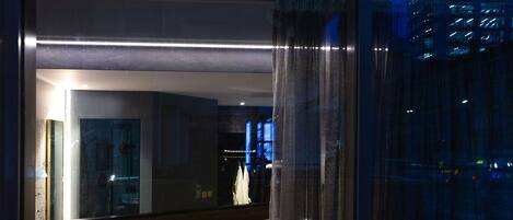A modern hotel room with a large bed, a bedside table with a lamp, and a view of the cityscape through large windows.