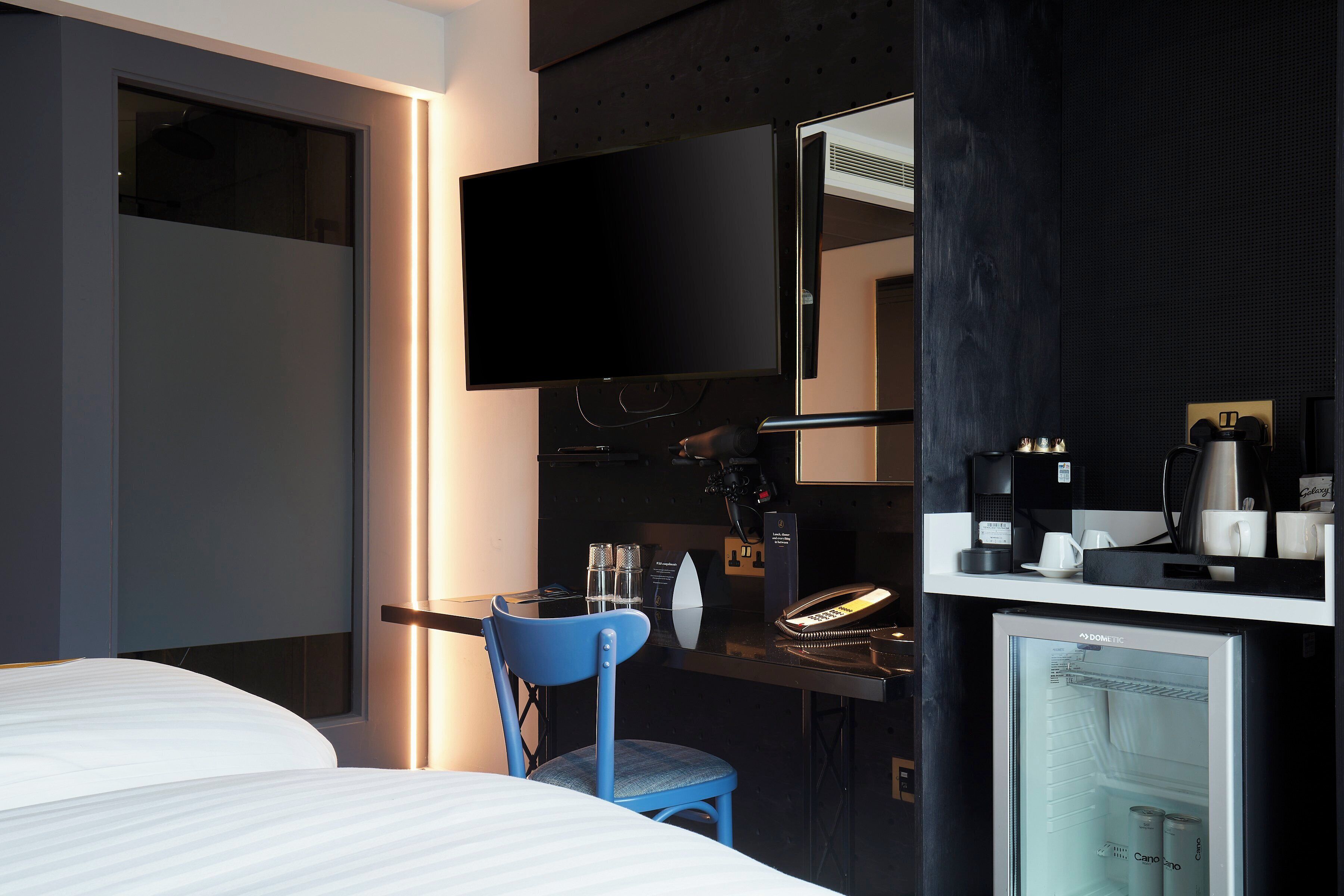 A hotel room with a bed, a desk with a computer, a chair, a TV, a mini-fridge, and a coffee machine.
