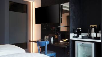 A hotel room with a bed, a desk with a computer, a chair, a TV, a mini-fridge, and a coffee machine.
