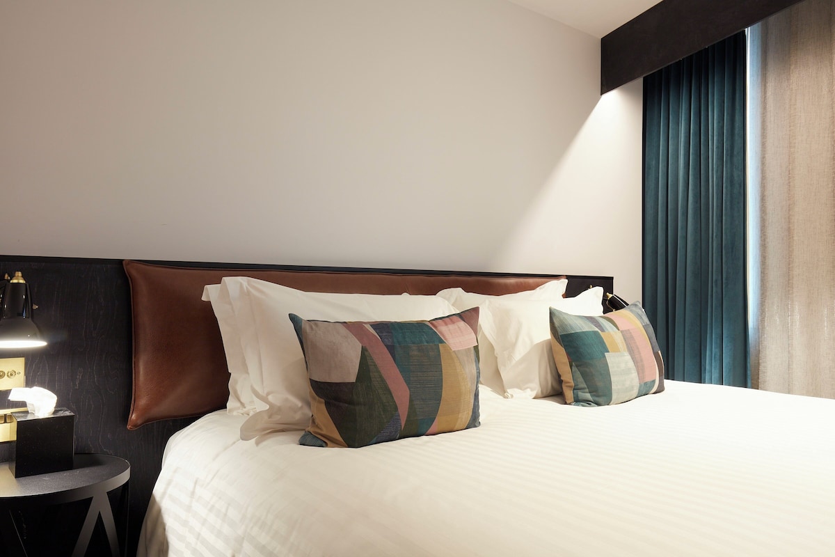A neatly made bed with multiple pillows, a bedside table with a lamp, and a window with curtains.