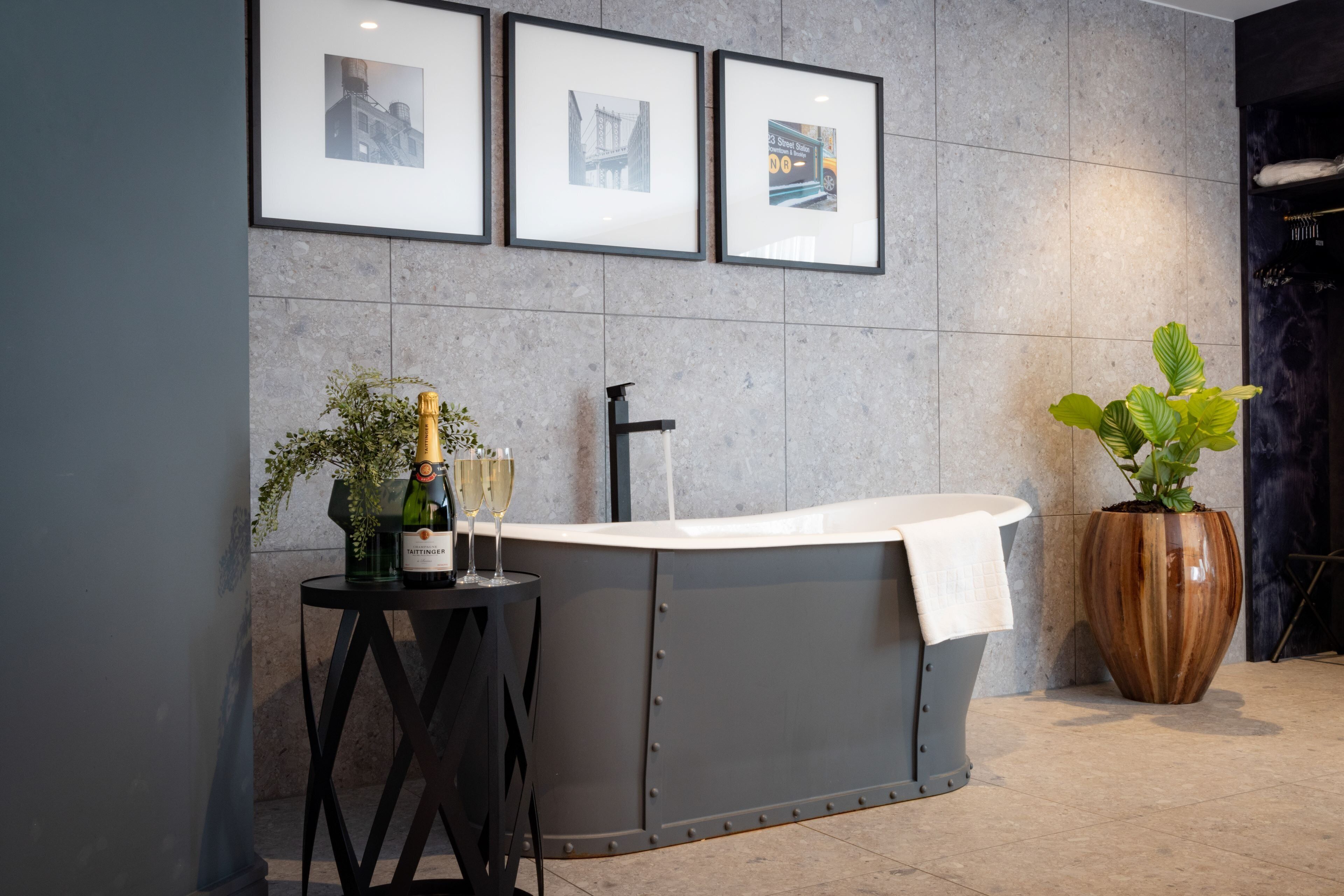 A modern bathroom with a freestanding bathtub, a small table with a bottle of champagne and glasses, and a large potted plant.
