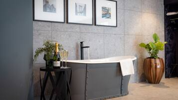 A modern bathroom with a freestanding bathtub, a small table with a bottle of champagne and glasses, and a large potted plant.