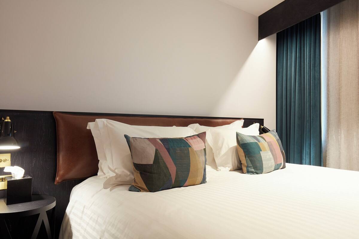 A neatly made bed with multiple pillows, a bedside table with a lamp, and a window with curtains.