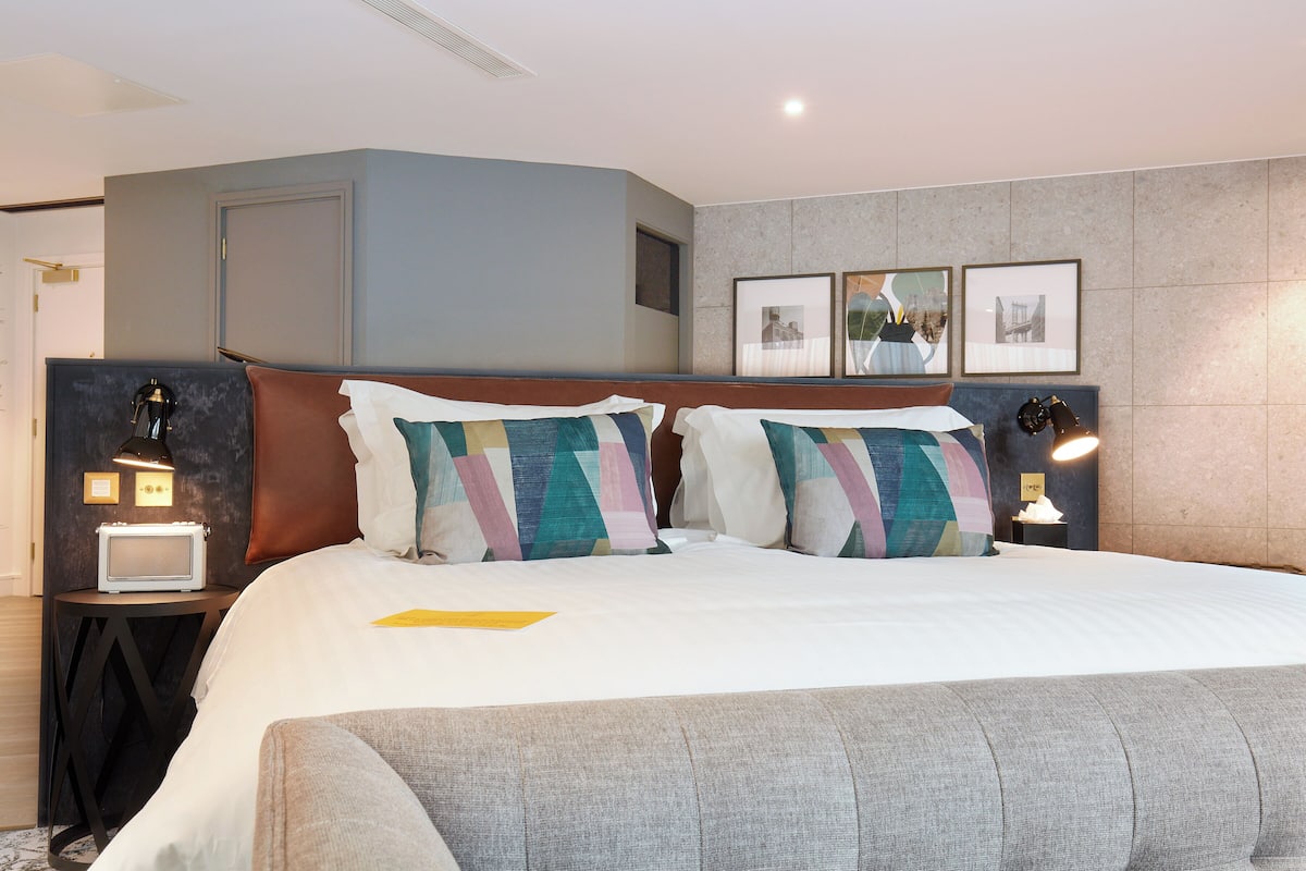 A modern hotel room with a large bed, two bedside tables, a nightstand, and a wall-mounted lamp.