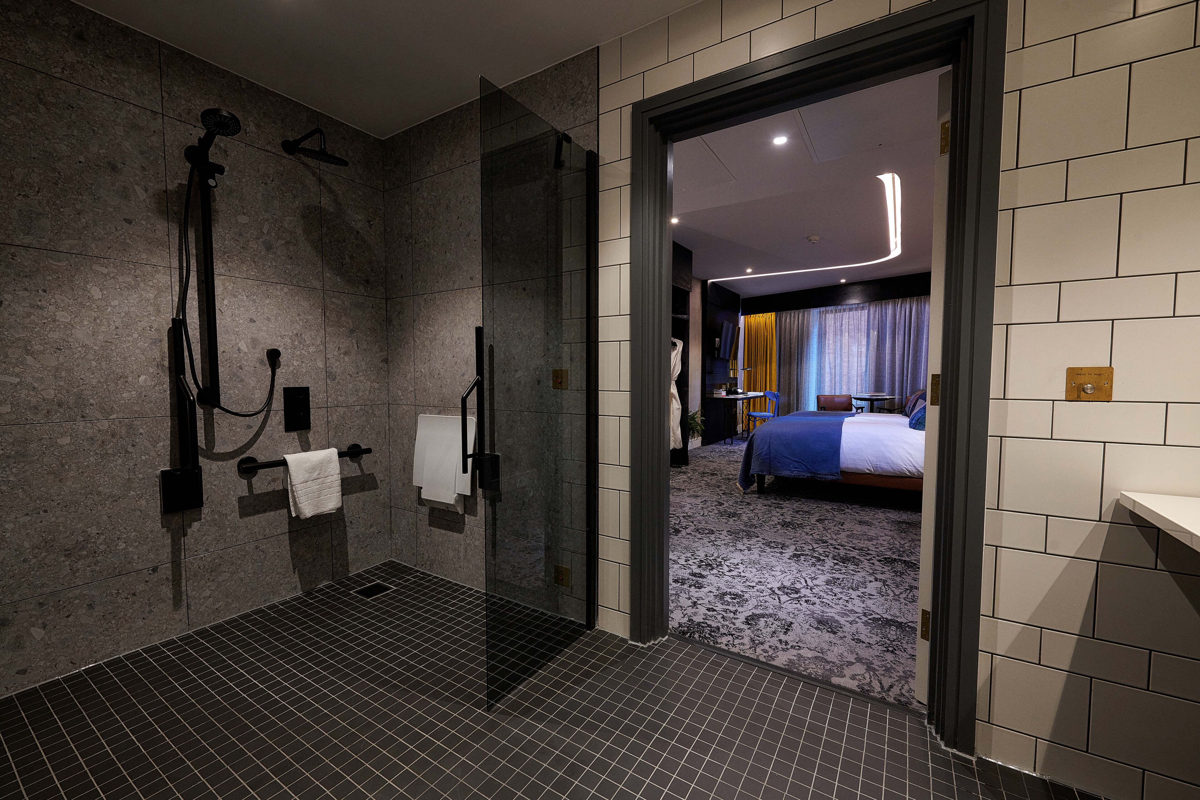 A modern bathroom with a glass shower enclosure, a towel rack, and a visible bedroom with a bed and curtains.