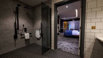 A modern bathroom with a glass shower enclosure, a towel rack, and a visible bedroom with a bed and curtains.