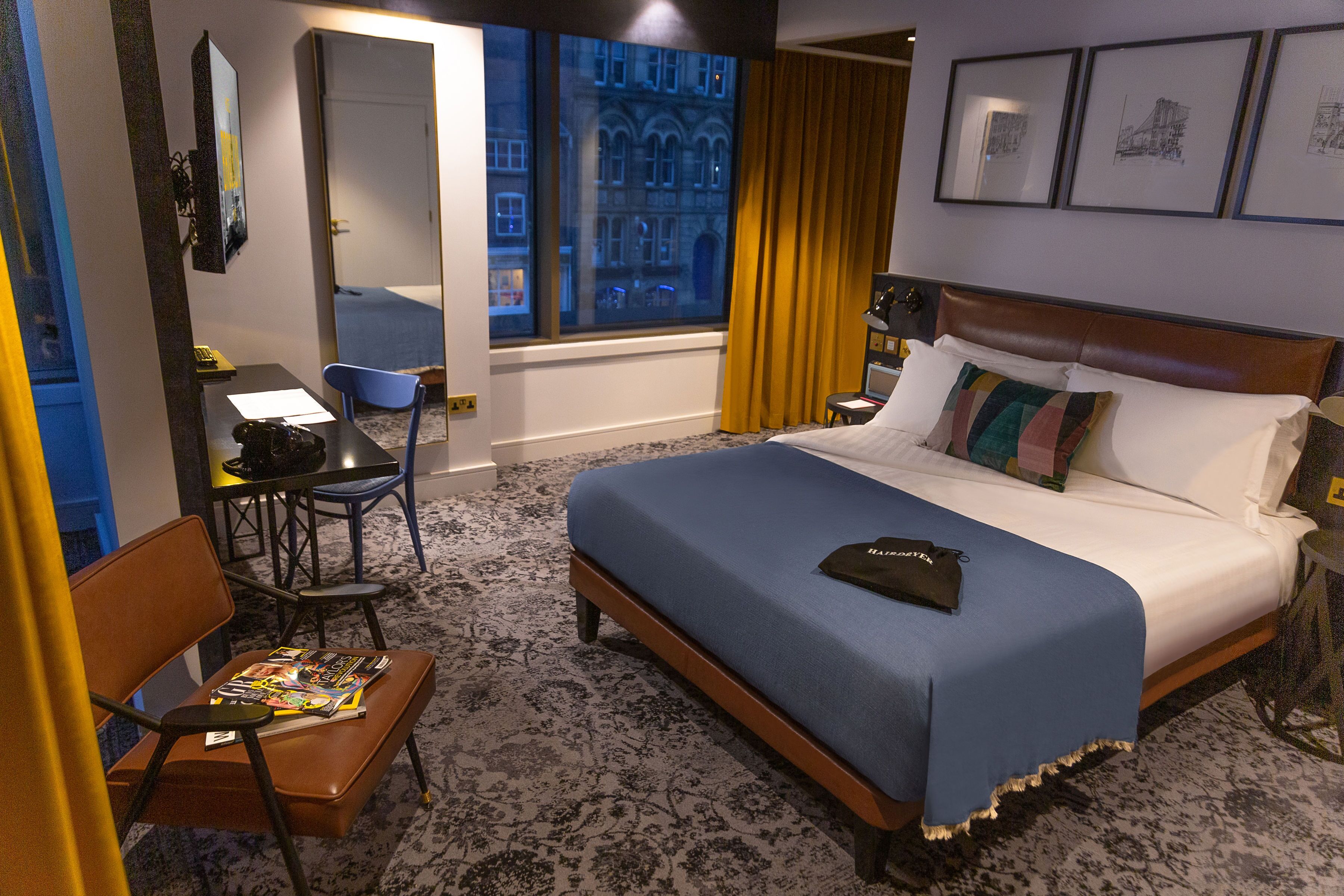 A hotel room with a large bed, a desk with a chair, a window with curtains, and framed pictures on the wall.