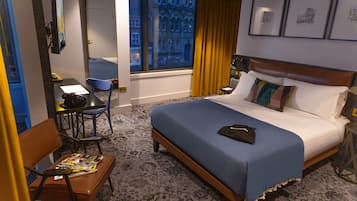 A hotel room with a large bed, a desk with a chair, a window with curtains, and framed pictures on the wall.