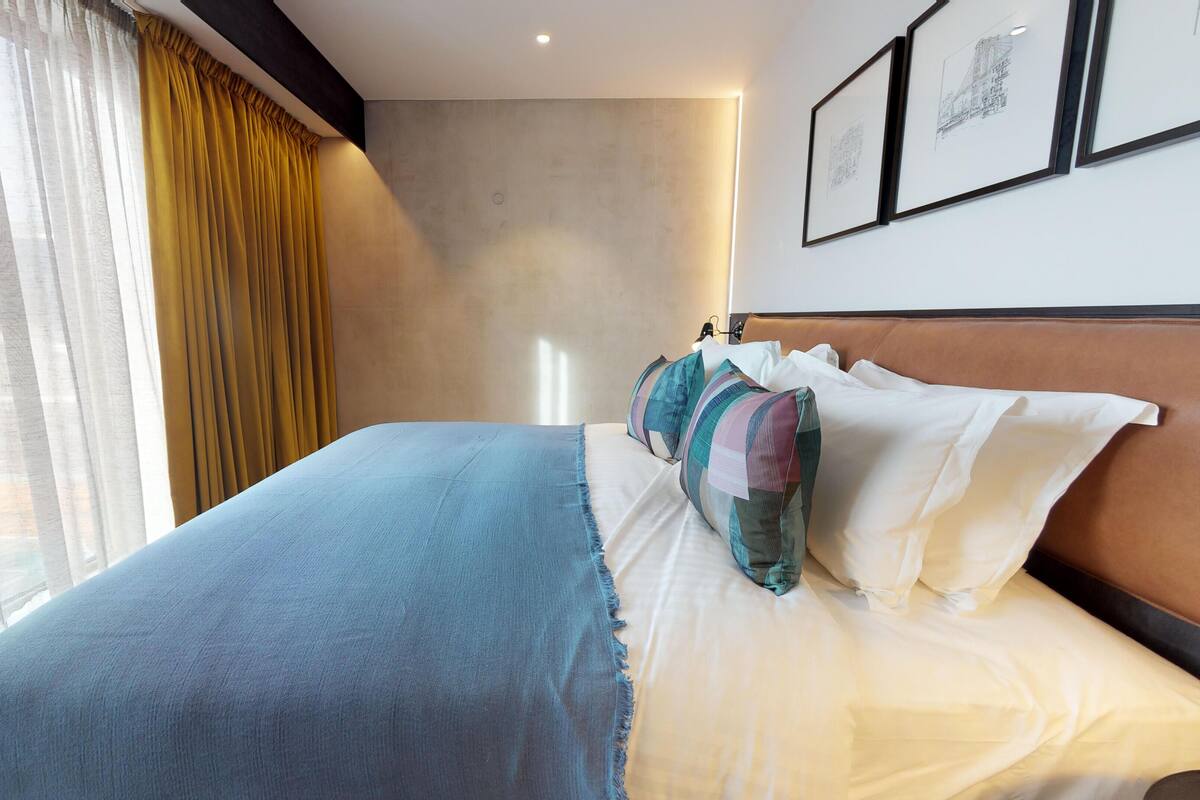 A hotel room with a large bed, a headboard, and a window with curtains. There are framed pictures on the wall.