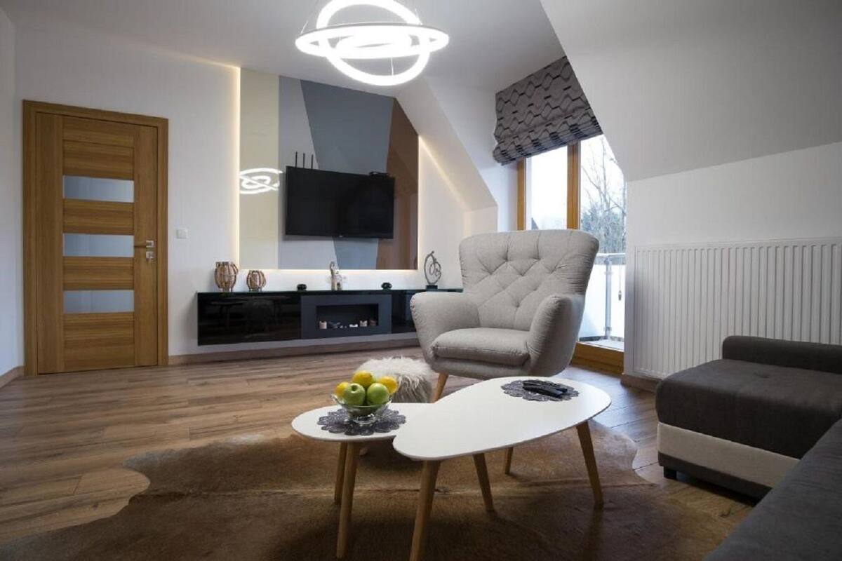 A modern living room with a large window, a flat-screen TV, a grey armchair, a white coffee table with fruit, and a dark sofa.