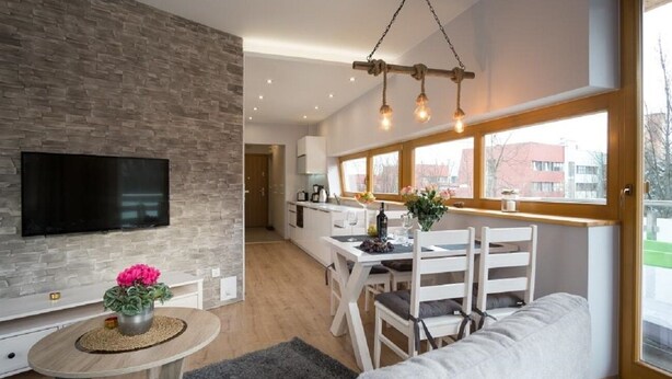 En moderne stue med et fladskærms-tv, et rundt sofabord med en vase, og en sofa. Der er et spiseområde med et bord og stole, og et køkken i baggrunden.