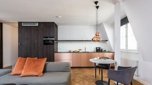 A modern living room with a grey sofa, a small round table, and a grey chair. There is a kitchen area with a copper pendant light and a window with a white blind. - Exclusive home in heart of Prague (Prague)