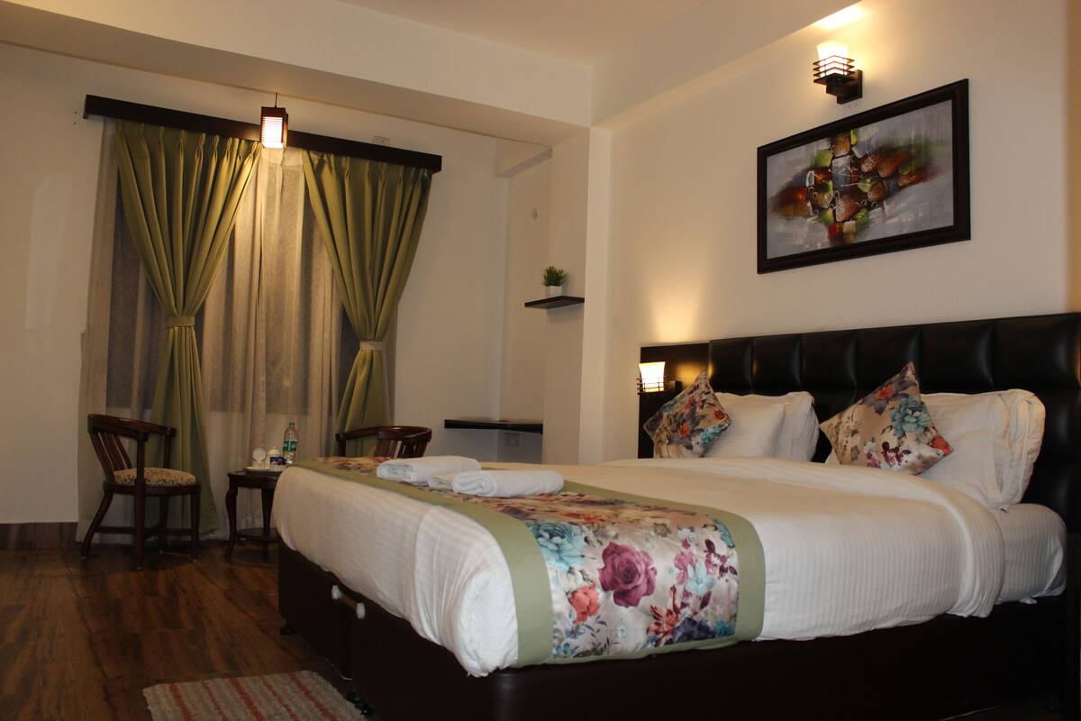 A hotel room with a bed, a chair, a small table, a nightstand, and a painting on the wall.