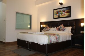 A neatly made bed with a floral bedspread, a large window with blinds, and a framed abstract painting on the wall. - The Leenor Regency (Gangtok)