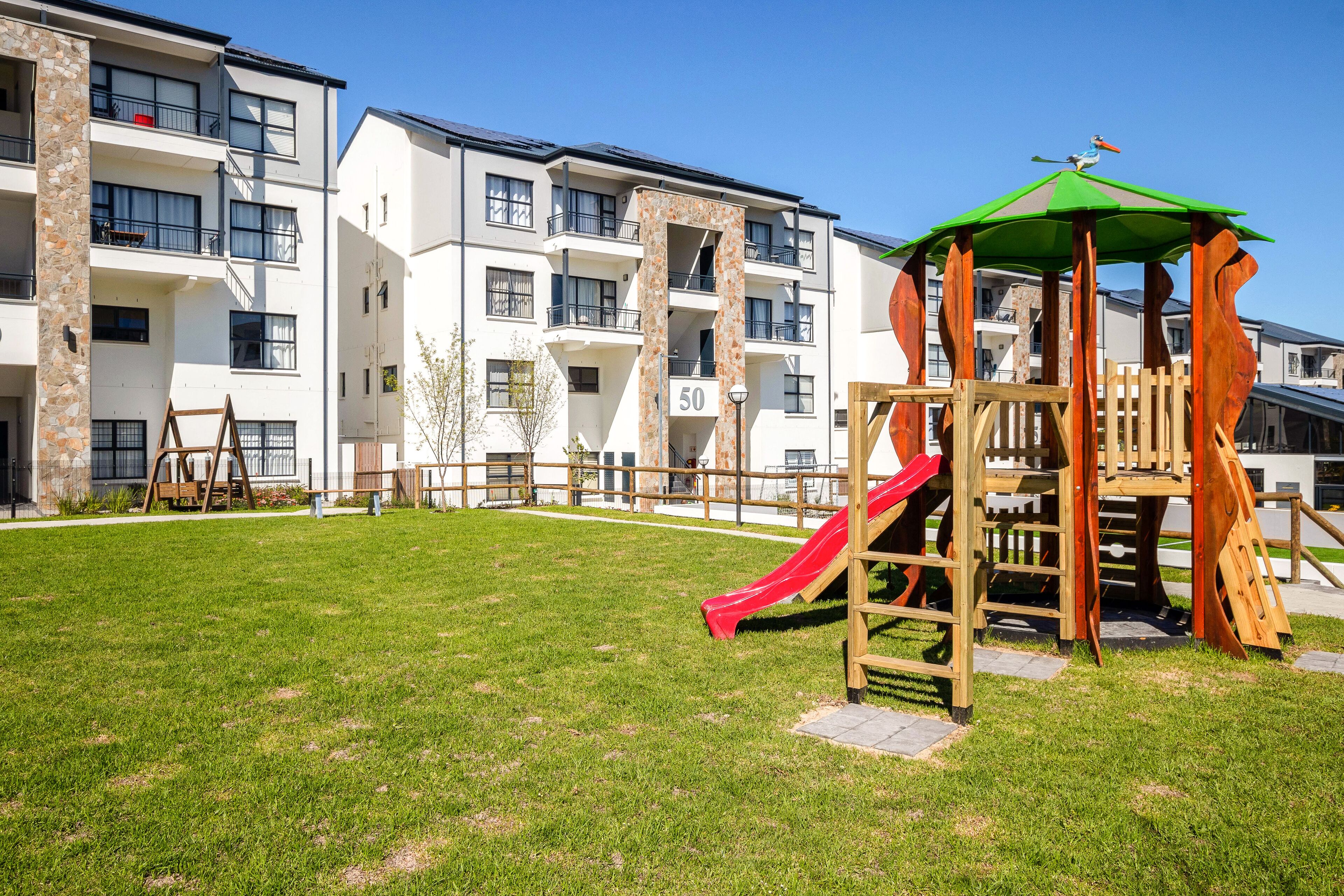A playground with a slide and climbing structures, surrounded by apartment buildings with balconies and a clear sky.