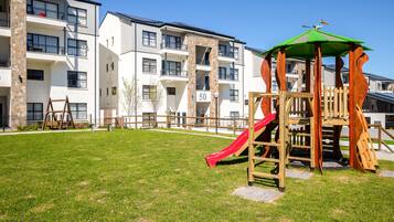 A playground with a slide and climbing structures, surrounded by apartment buildings with balconies and a clear sky.