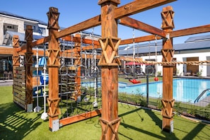Een houten speelconstructie met touwen en een zwembad op de achtergrond.