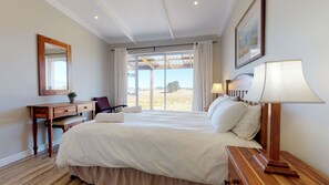 A bedroom with a large bed, a wooden desk, a chair, a lamp, and a view of the outdoors.