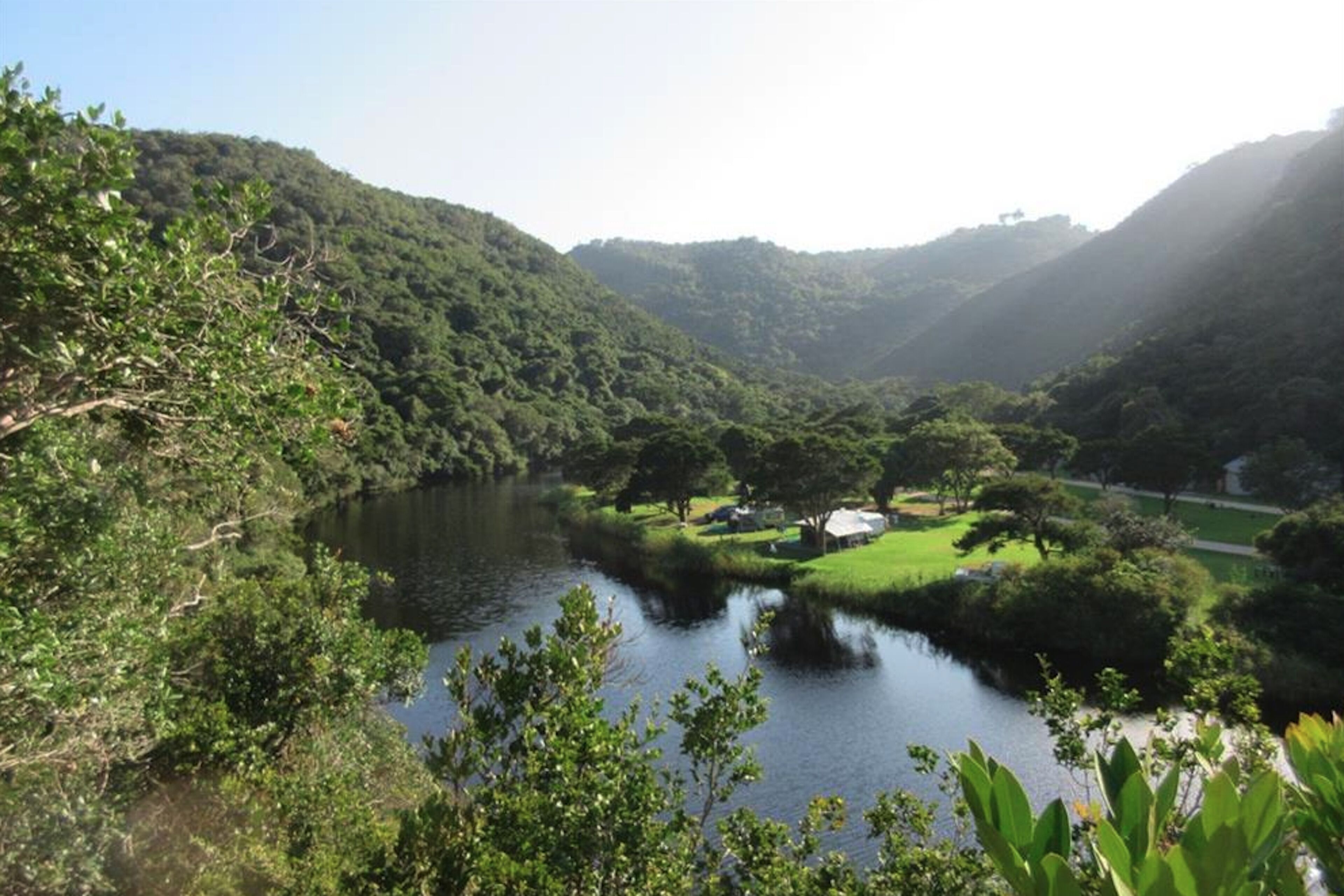A serene landscape with a river, lush greenery, and a small building near the water.