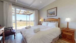 A hotel room with a large bed, two bedside lamps, a desk with a chair, and a view of a landscape through a sliding glass door. - Aleph Cottage George (George)