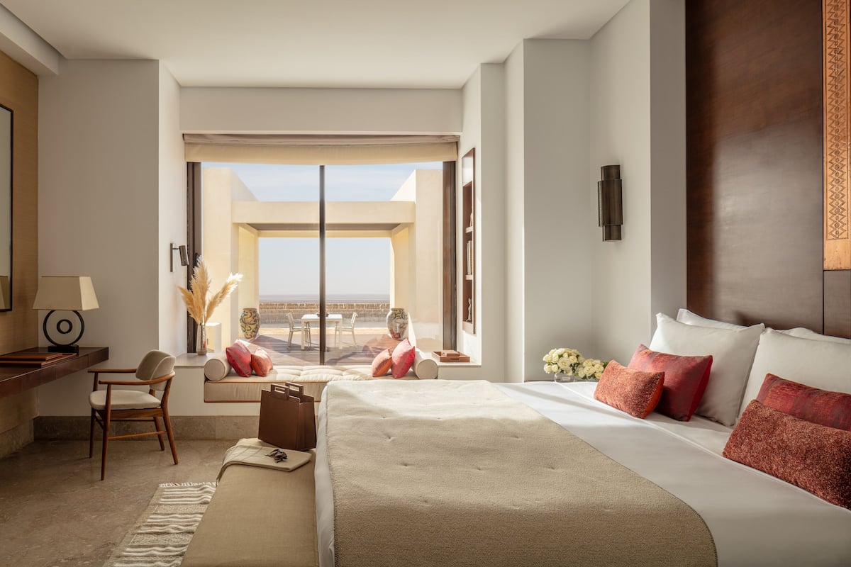 A hotel room with a large bed, a desk with a lamp, a chair, and a view of the outdoors.