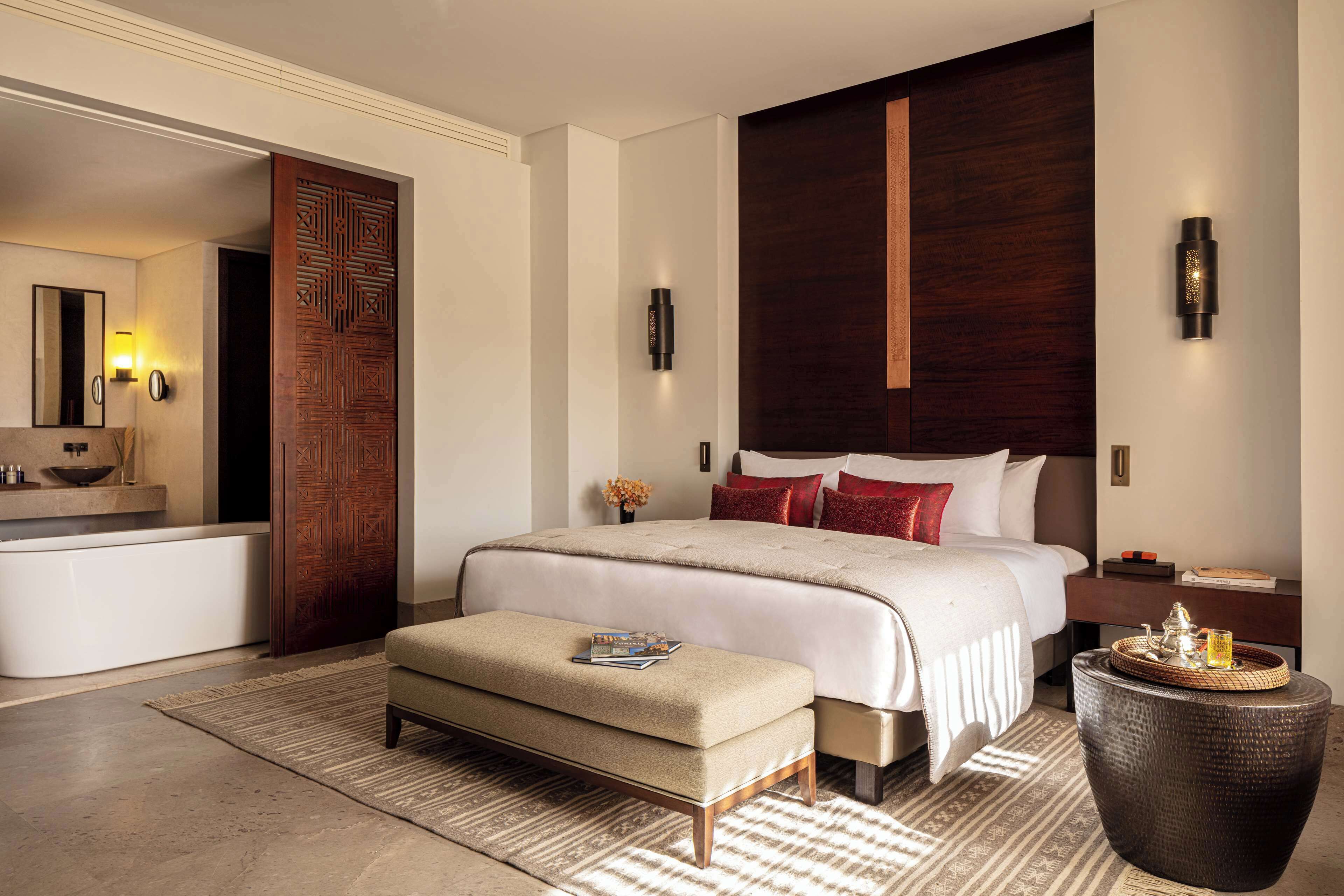 Une chambre d’hôtel moderne avec un grand lit, une table de chevet, une commode, une porte en bois, une salle de bain avec baignoire et une grande plante en pot.