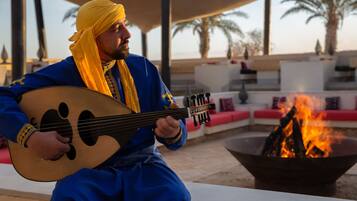 A person playing a stringed instrument outdoors, seated at a table with a fire pit in the background.