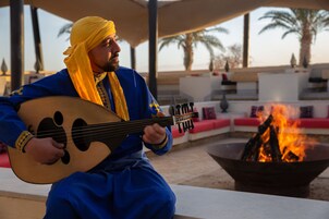 A person playing a stringed instrument outdoors, seated at a table with a fire pit in the background.