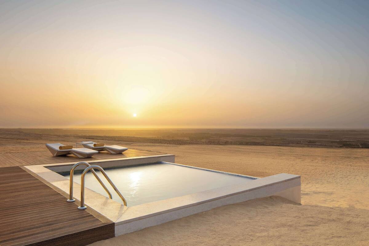 A sunlit infinity pool with lounge chairs and a wooden deck, overlooking a vast desert landscape.