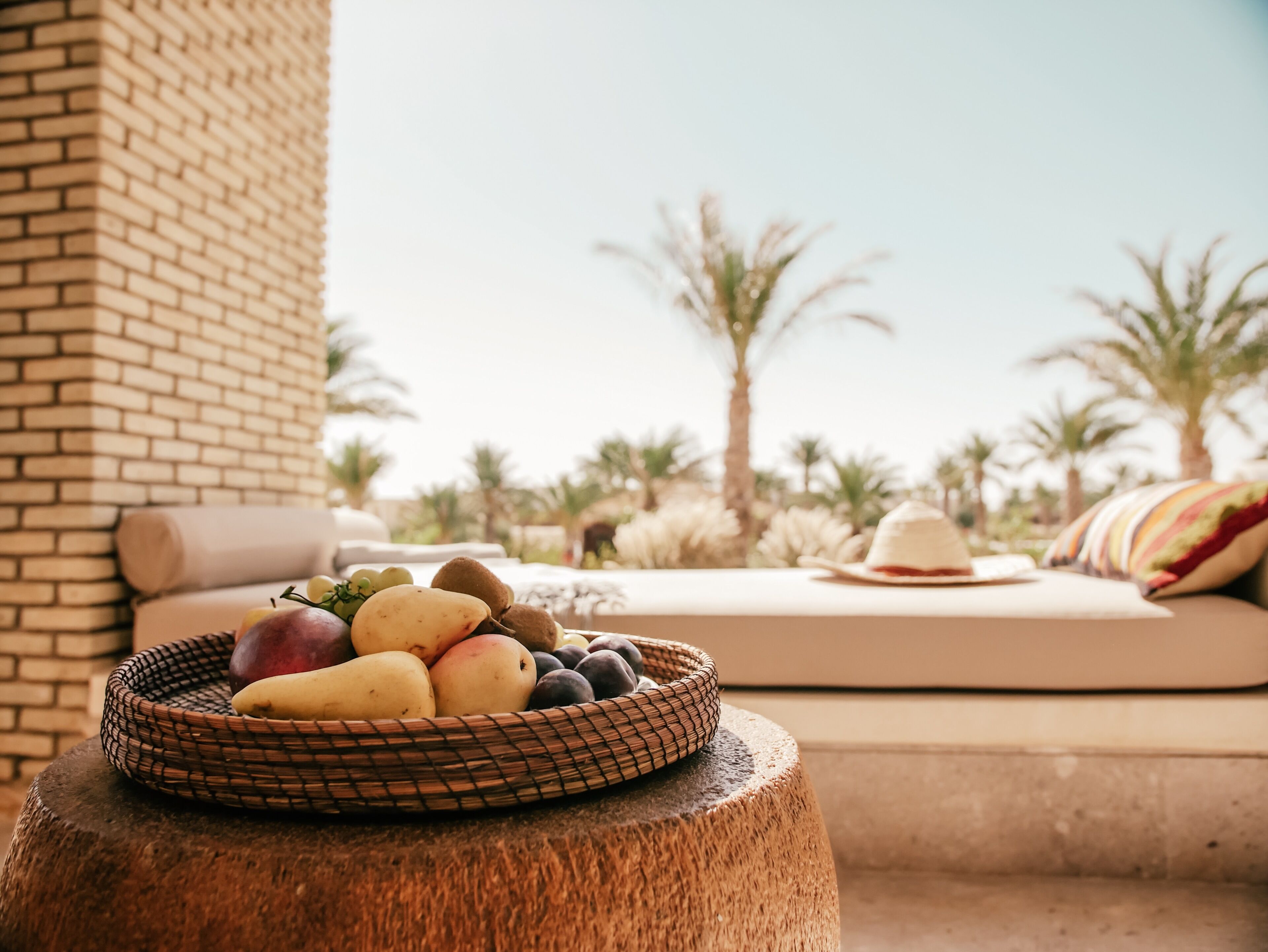 A wicker basket of fruits on a wooden stool, a cushioned outdoor seating area with a hat, and palm trees in the background.