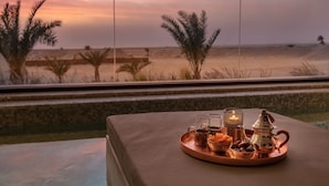 A sunset view from a window with a tray of drinks and snacks on a cushioned surface. - The Mora Sahara Tozeur (Tozeur)