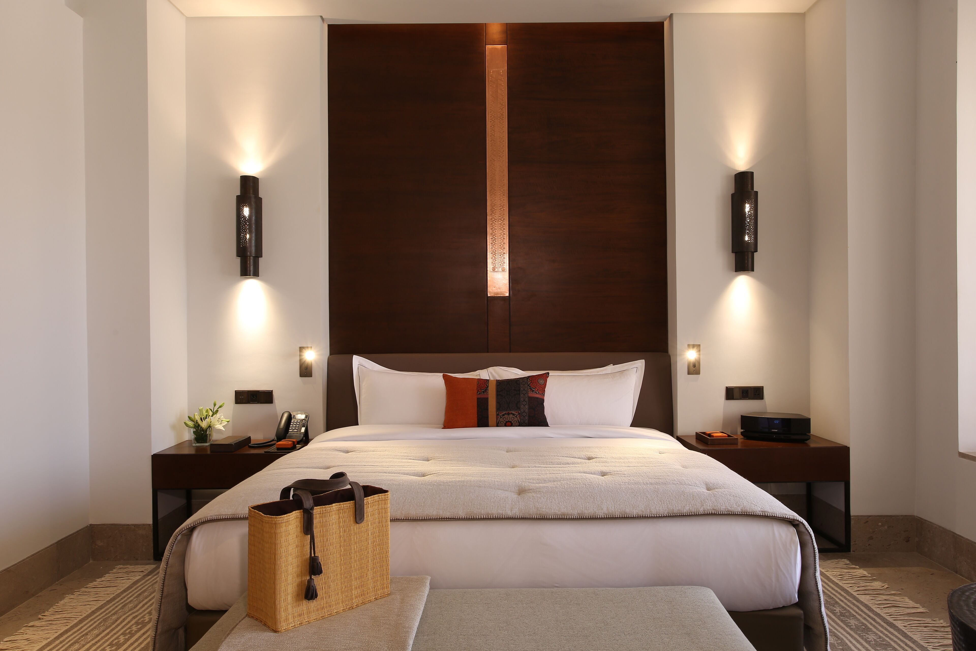 Una camera d'albergo moderna con un letto grande, due comodini, un tavolino e un cesto di vimini, oltre a una lampada a parete.