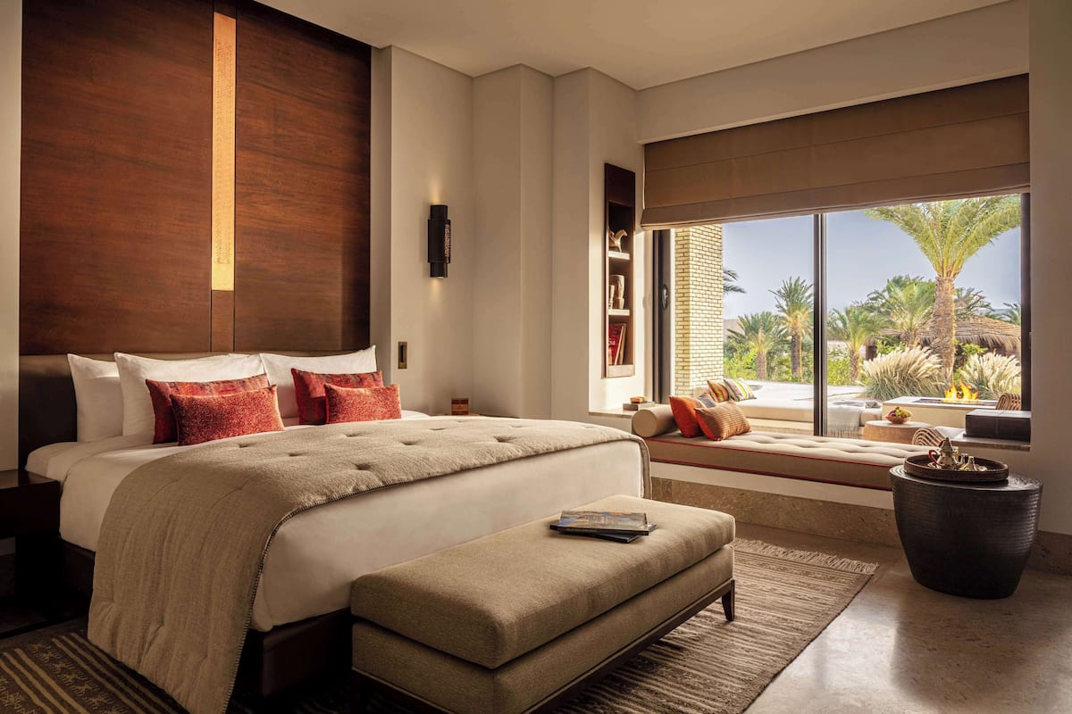 A modern bedroom with a large bed, a bench, a large window with a view of palm trees and a brick wall.