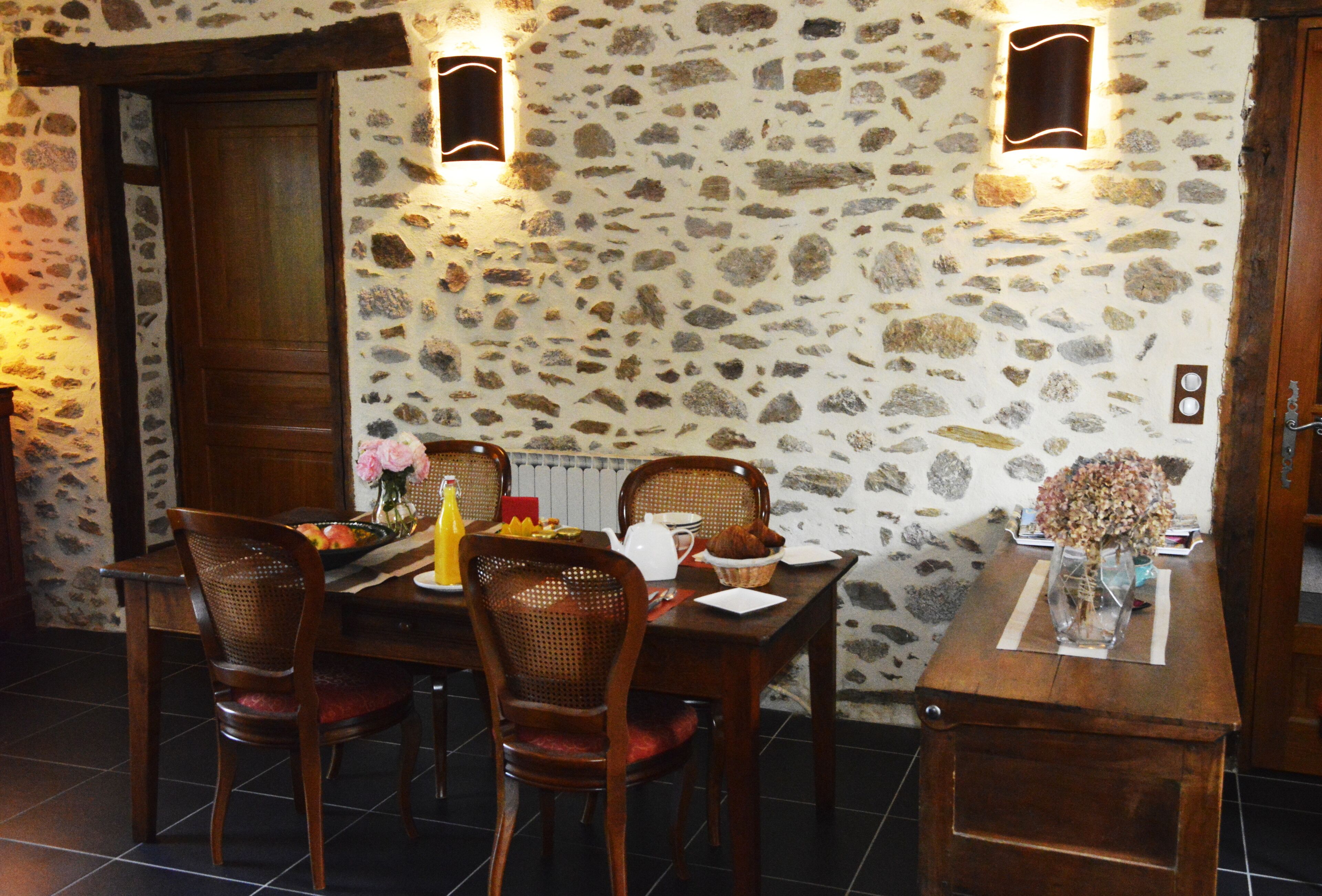 Zona pranzo con parete in pietra, tavolo in legno, sedie e un piccolo tavolino con un vaso.