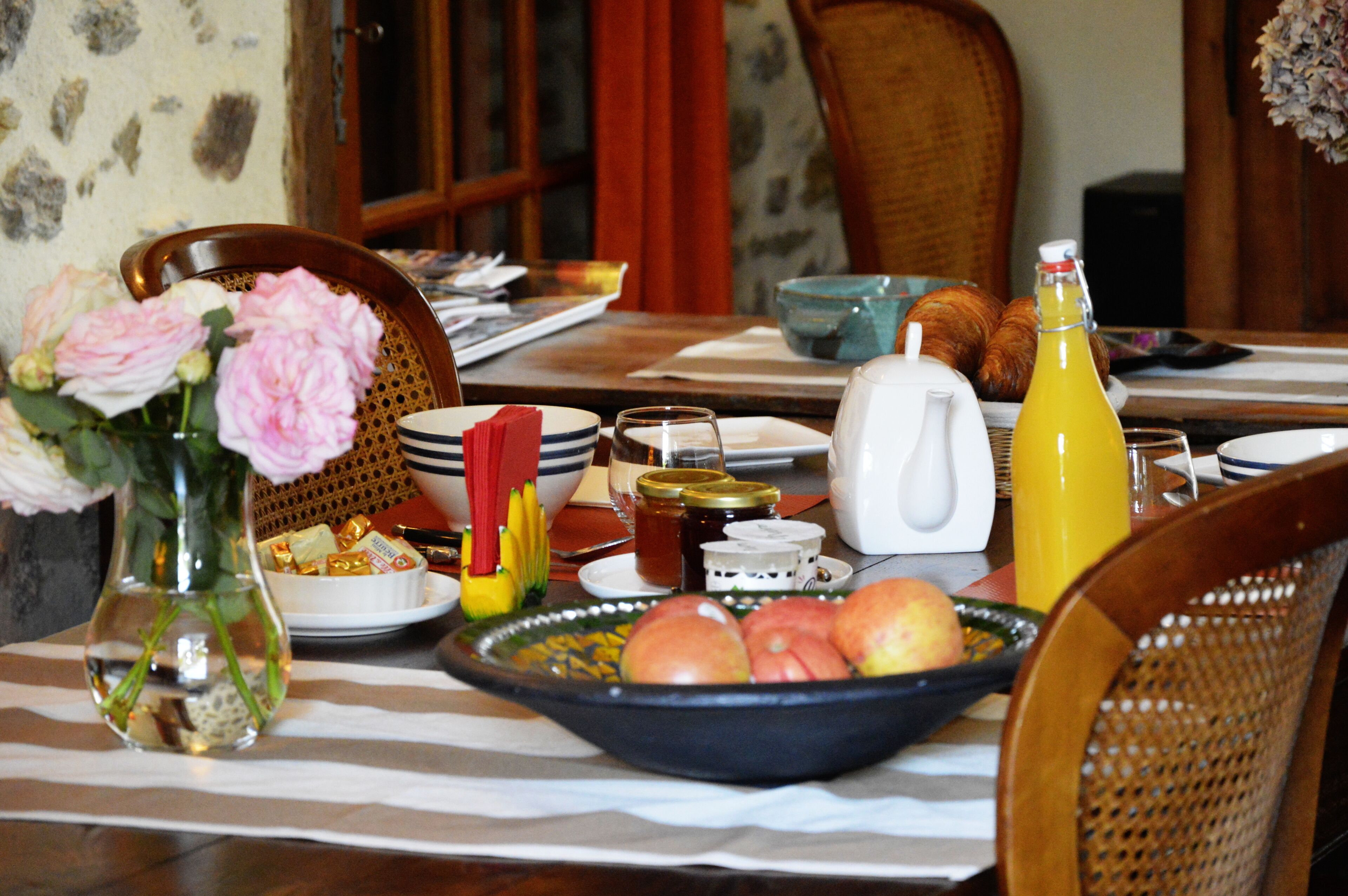A dining table set with breakfast items including fruits, a bottle of juice, and a vase with flowers.
