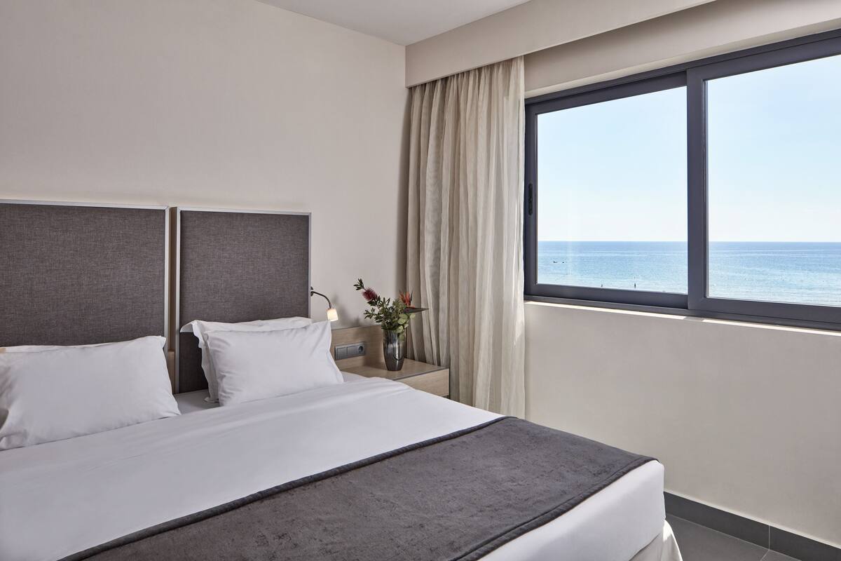 A hotel room with a large bed, a nightstand with a lamp, and a view of the sea through a window.