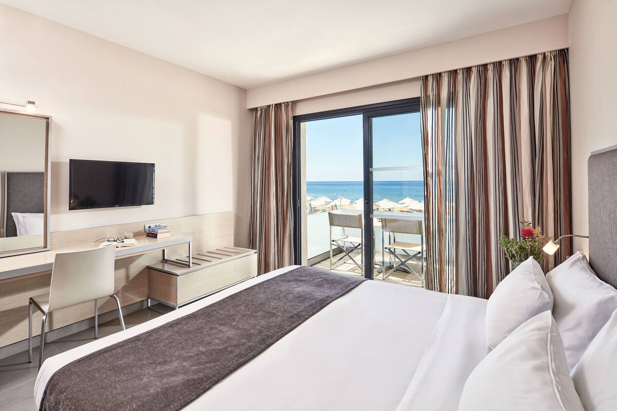 A hotel room with a bed, a desk, a chair, a TV, and a view of the beach.