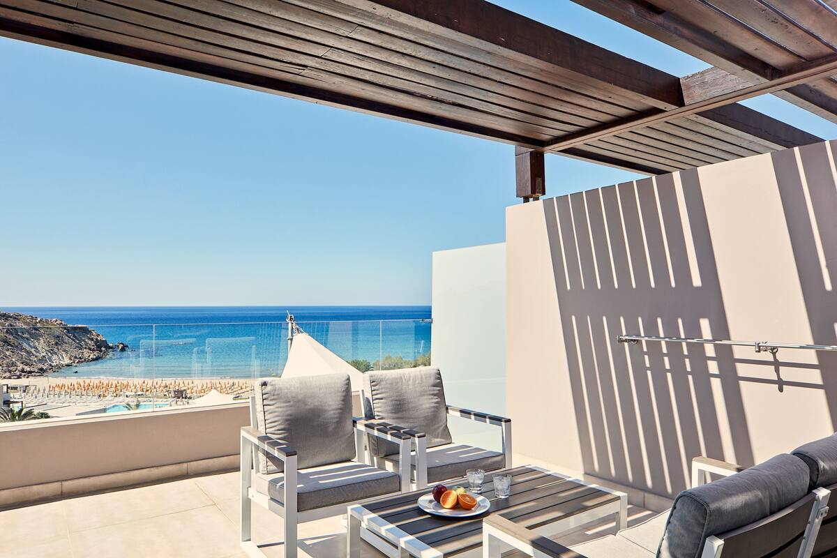 A balcony with a view of the sea, a table with a plate of fruit, and two chairs.