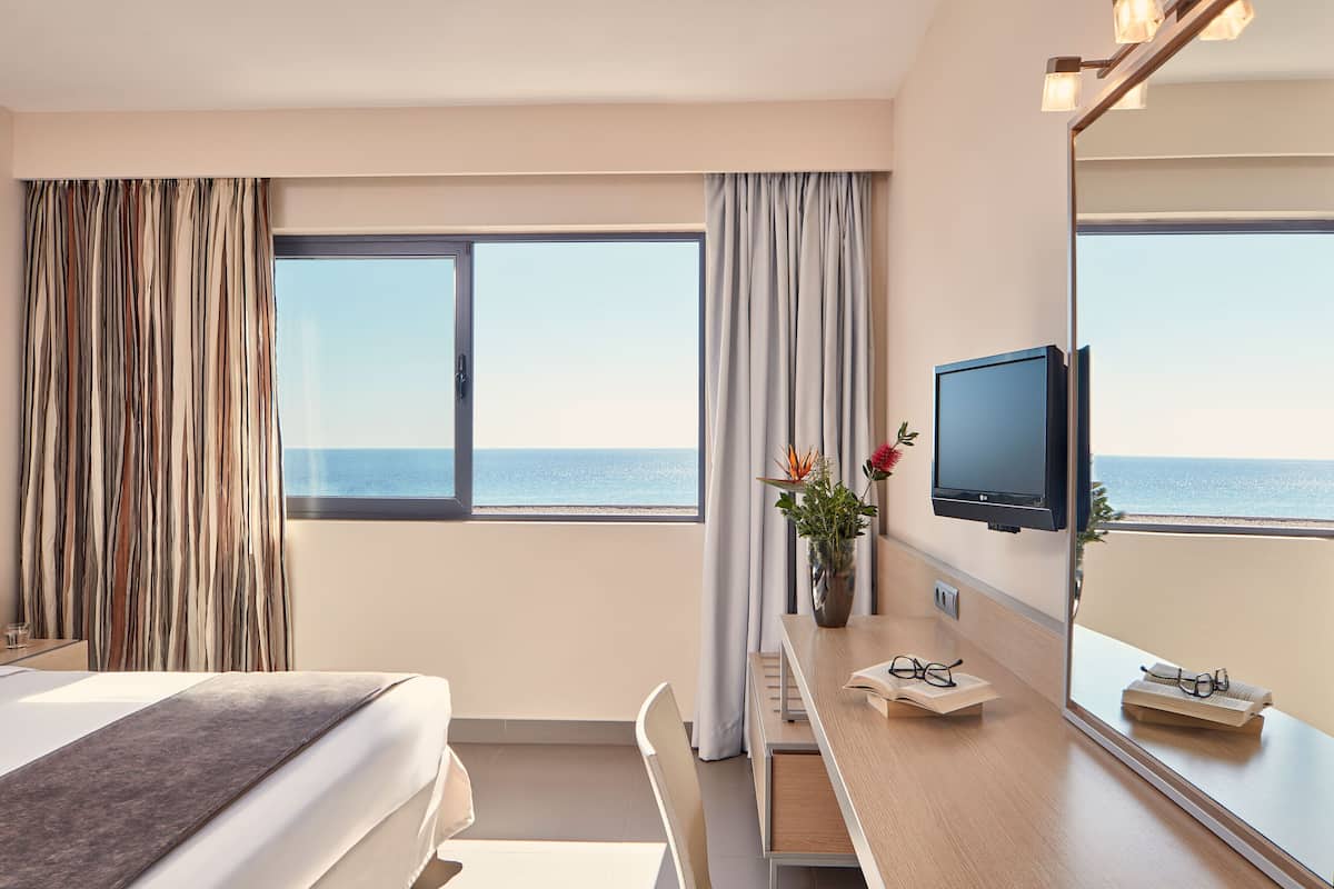A hotel room with a bed, a desk with a chair, a television, a large window with a view of the sea, and a mirror.
