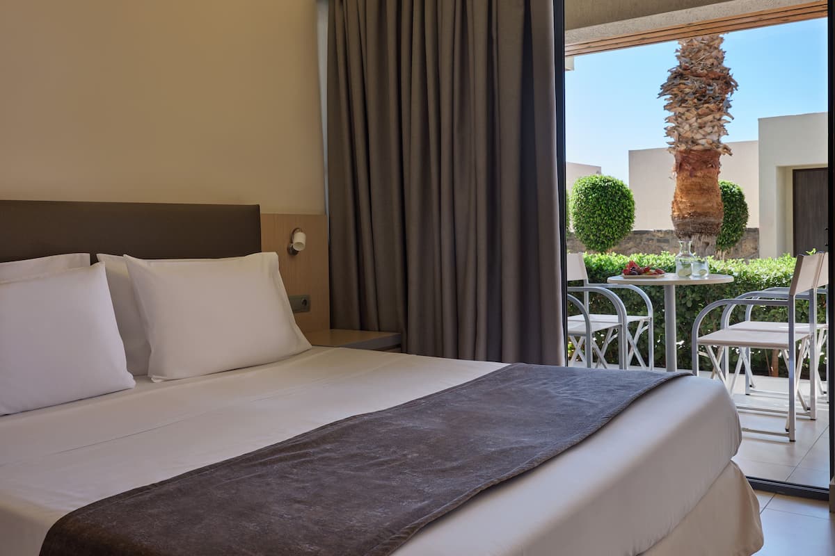 A hotel room with a bed, a nightstand, a window with curtains, and a view of an outdoor area with a table and chairs.
