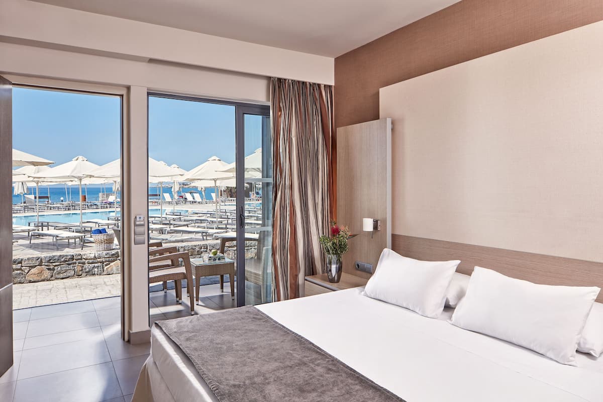 A hotel room with a large bed, a view of a pool and umbrellas, and a balcony with a wicker table and chairs.