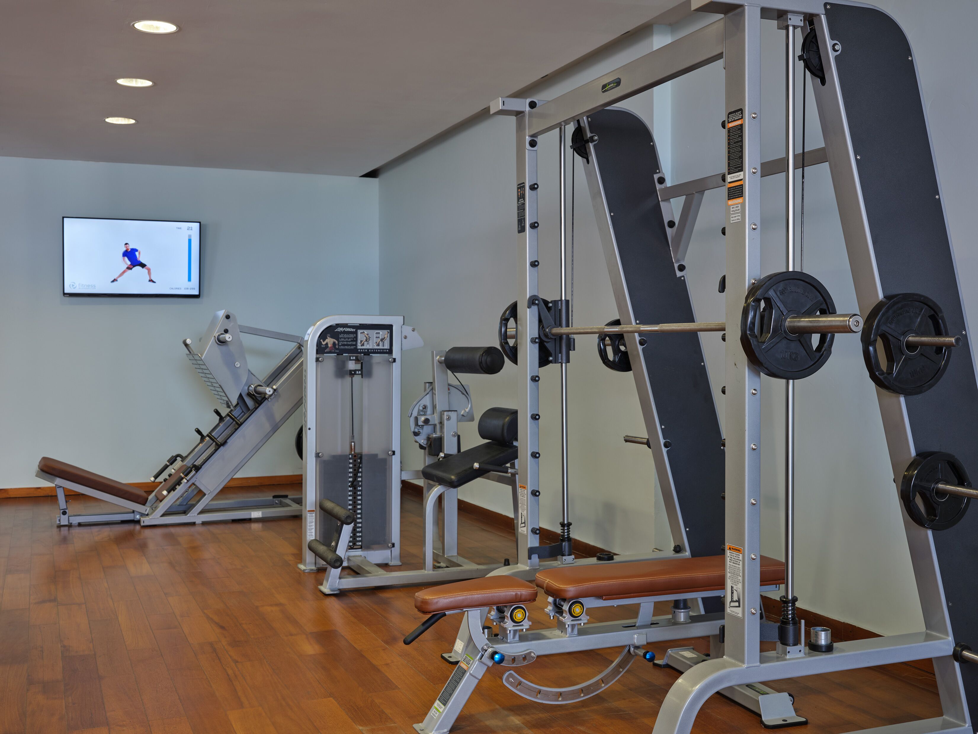 A gym with various exercise machines, a television displaying a workout video, and wooden flooring.