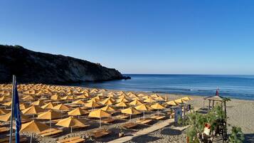 A beach with numerous yellow umbrellas and sun loungers, a wooden deck, and a rocky cliff in the background.